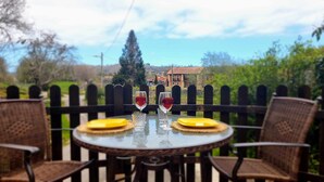 A table with two glasses of red wine, two yellow plates, and wicker chairs on a balcony with a view of a house and trees. - Los Gemelos Vacation Home (San Roque Del Acebal)