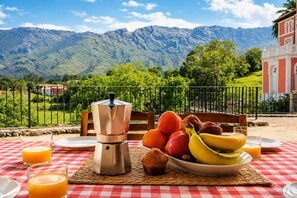En frukostuppsättning med en kaffebryggare, frukt och juice på ett rutigt bordsduk, med utsikt över bergen.
