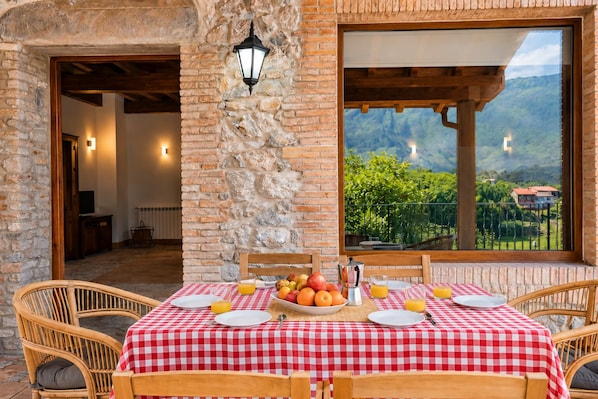 Un comedor con mantel a cuadros, sillas de mimbre, pared de piedra y vistas a montañas a través de un amplio ventanal.