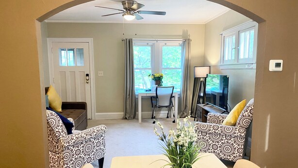 A living room with patterned sofas, a dining table, a ceiling fan, a TV, and a window with curtains.