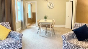 A living room with patterned sofas, a dining table with chairs, a clock on the wall, and a door leading to another room.