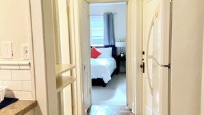 A narrow hallway leading to a bedroom with a bed, a nightstand, and a window with curtains.