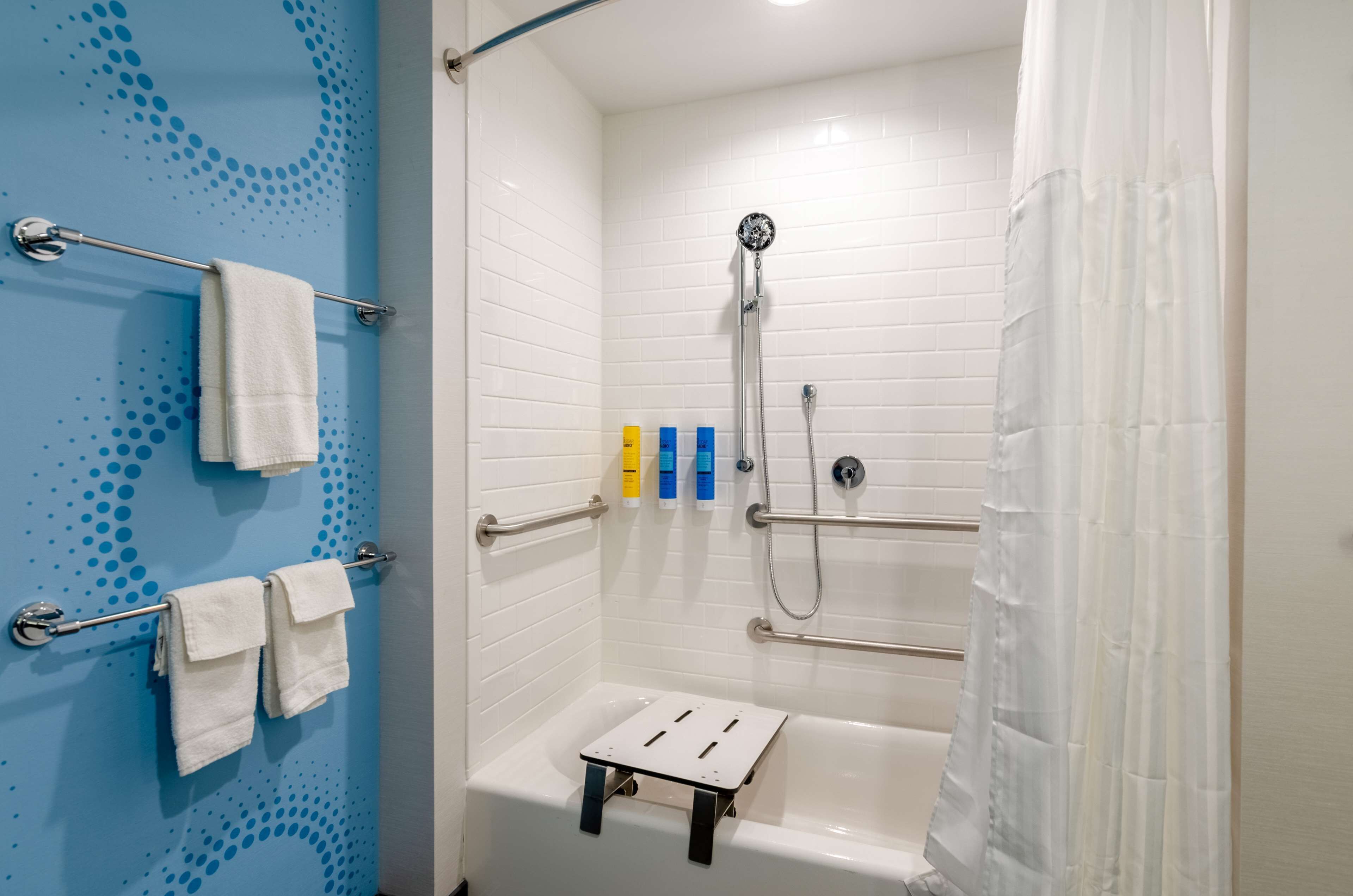 Et bruseområde med hvide flisevægge, brusearmatur, håndbruser og gelænder. Der er håndklæder på en holder og toiletartikler på væggen.