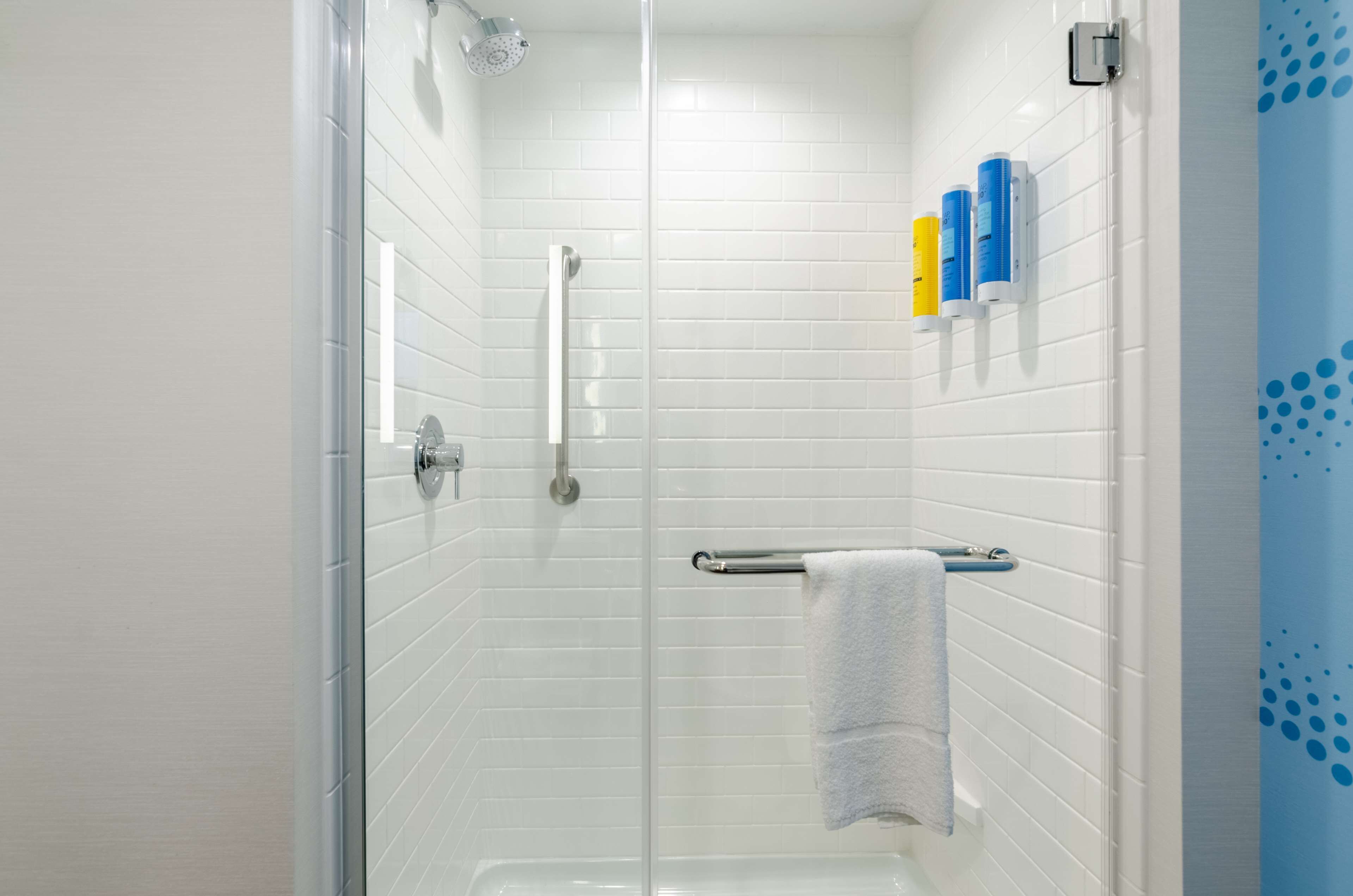 Un baño moderno con paredes de azulejos blancos, una cabina de ducha de vidrio, un cabezal de ducha manual, un perchero con una toalla blanca y una cortina de ducha azul.