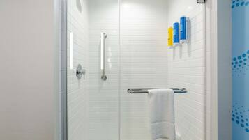 Un baño moderno con paredes de azulejos blancos, una cabina de ducha de vidrio, un cabezal de ducha manual, un perchero con una toalla blanca y una cortina de ducha azul.
