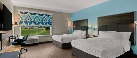 A hotel room with two beds, a desk, a chair, a TV, and a window with a view of a green lawn.