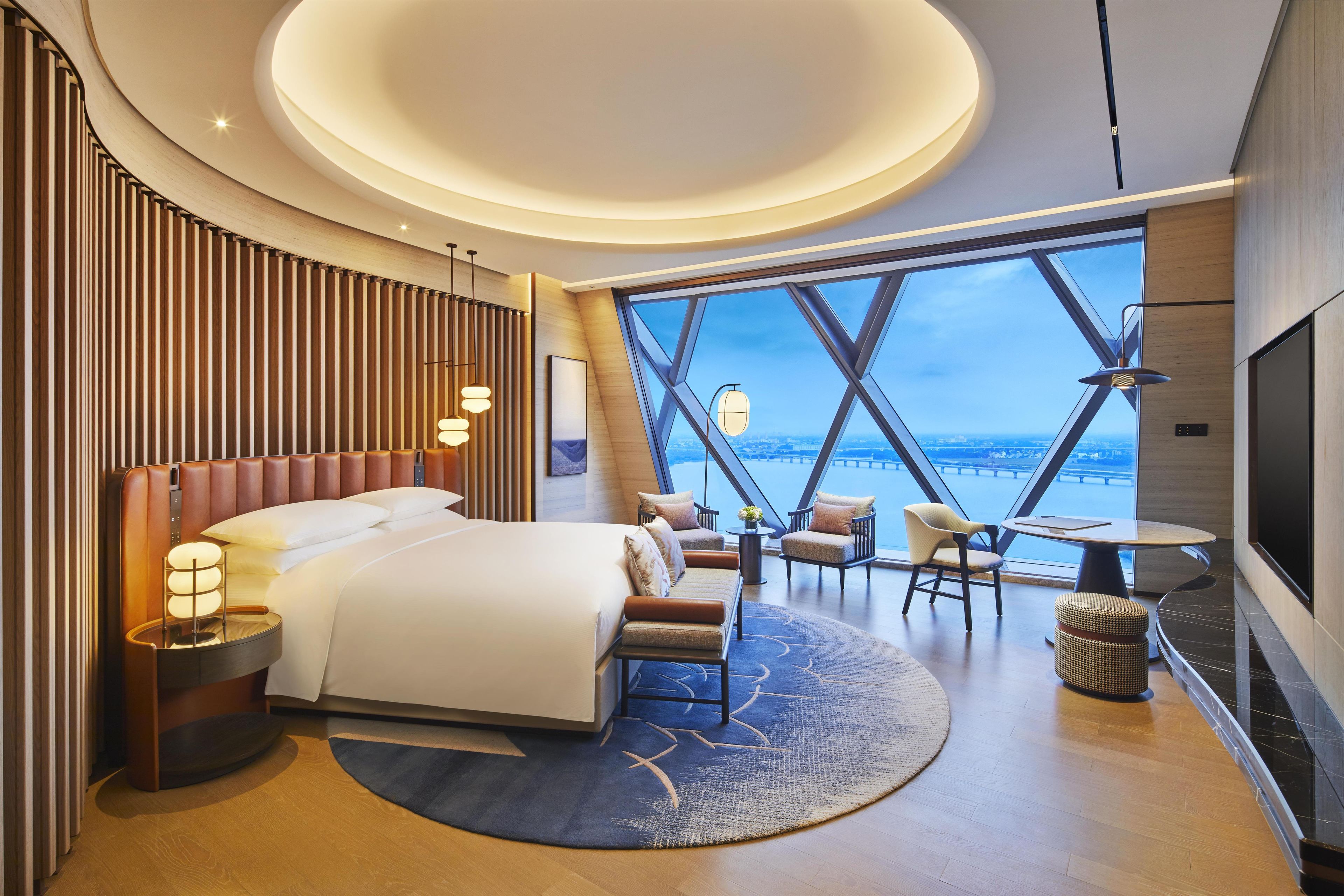 A modern hotel room with a large bed, seating area, and a view of the cityscape through large windows.