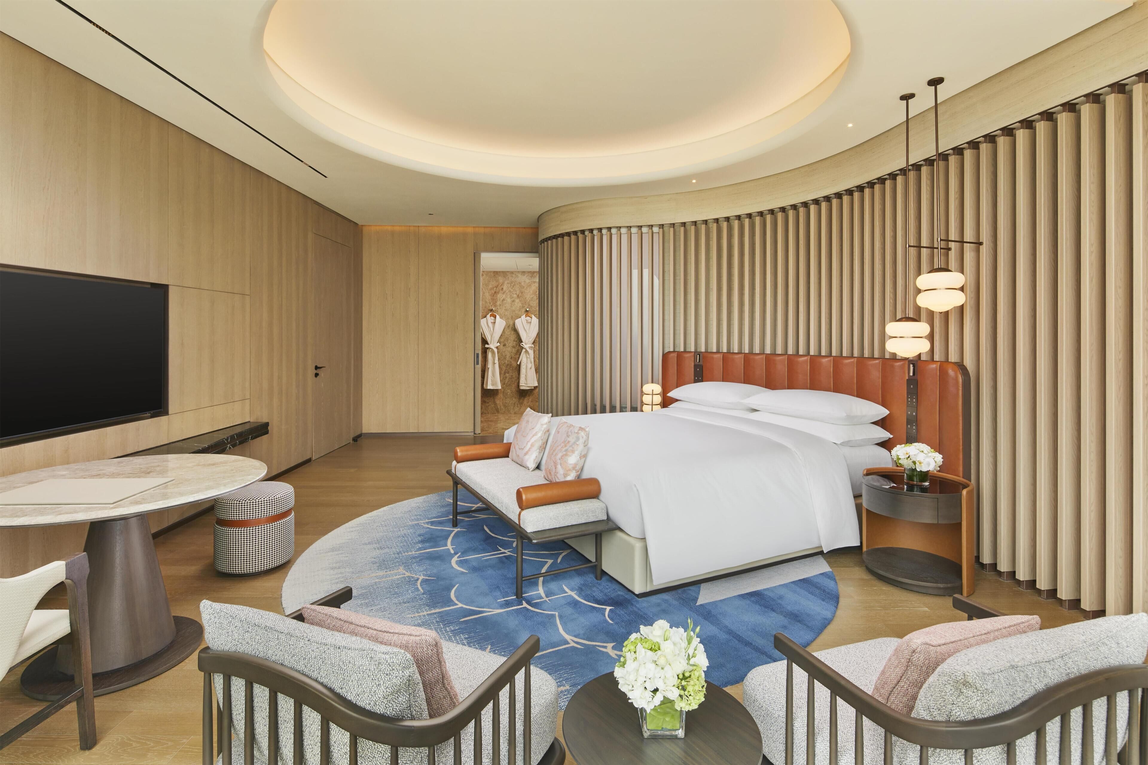 A modern hotel room with a large bed, a TV, a dining table, and a seating area.