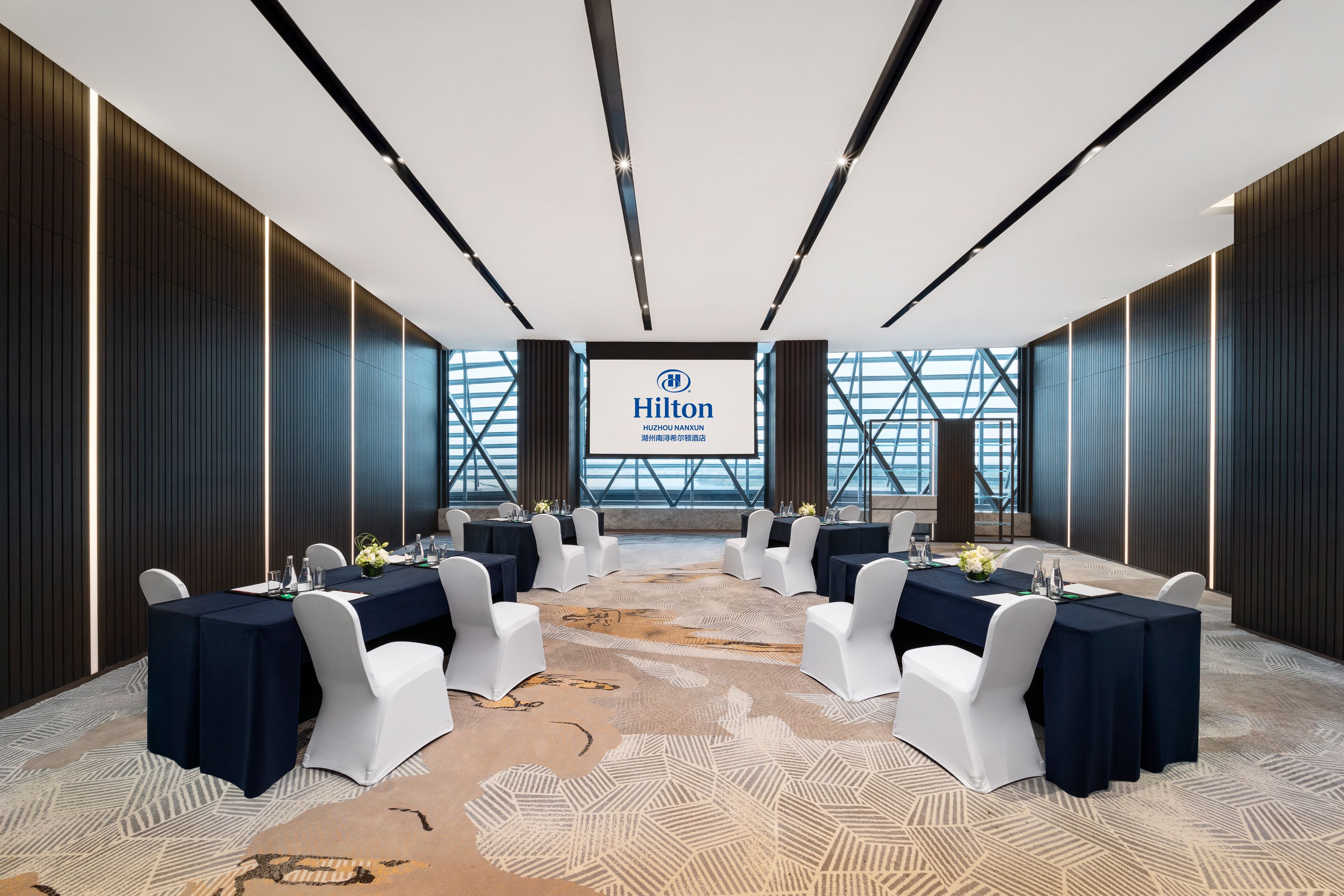 Sala de conferências com uma tela grande exibindo o logotipo da Hilton, cadeiras brancas e mesas preparadas para uma reunião.