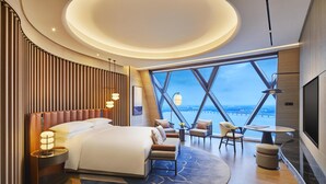 Een moderne hotelkamer met een groot bed, zithoek en uitzicht op de stad door grote ramen.
