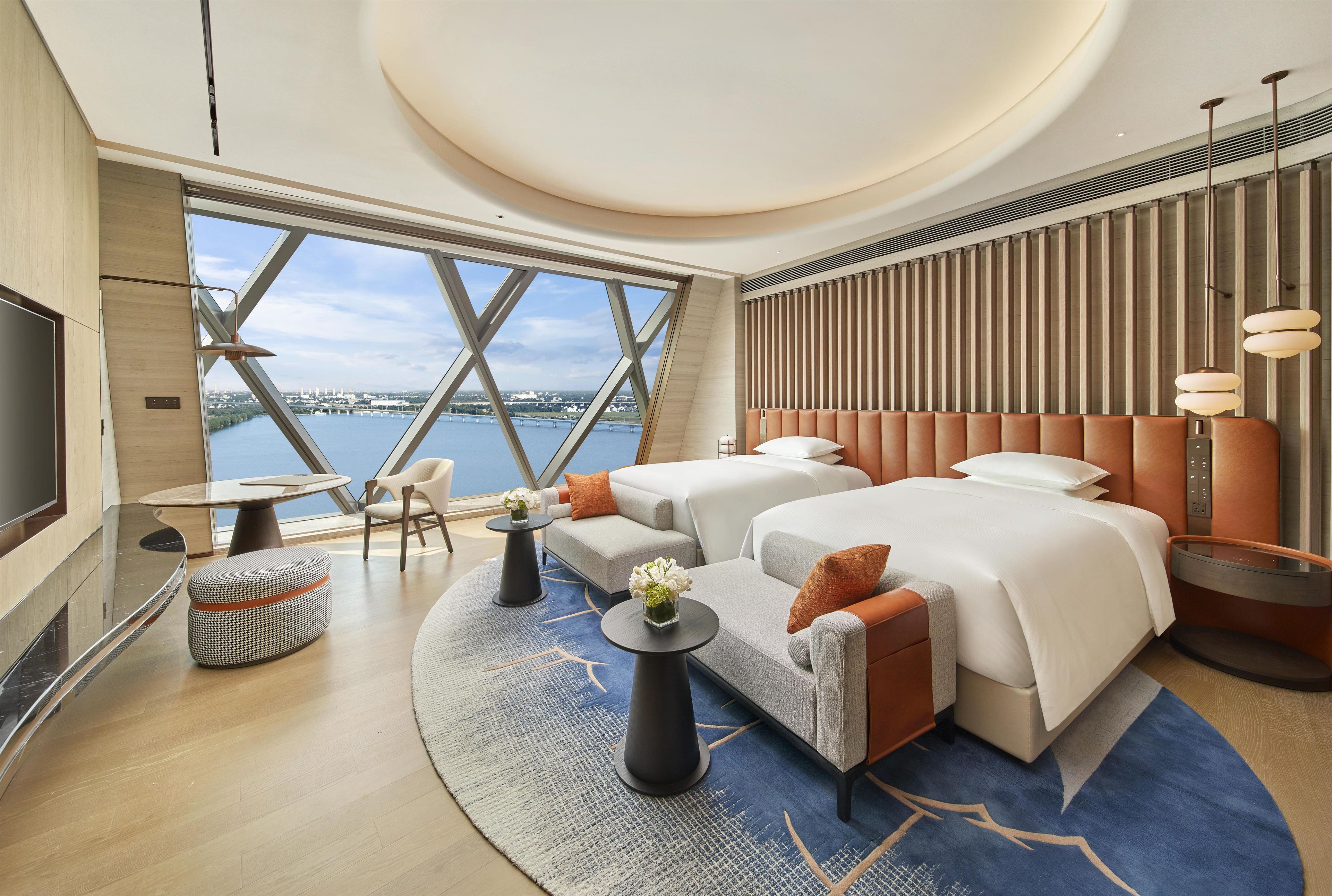 A modern hotel room with a large window, a bed, a sofa, a TV, and a dining area.