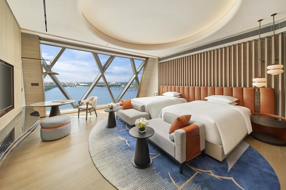 A modern hotel room with a large window, a bed, a sofa, a TV, and a dining area.