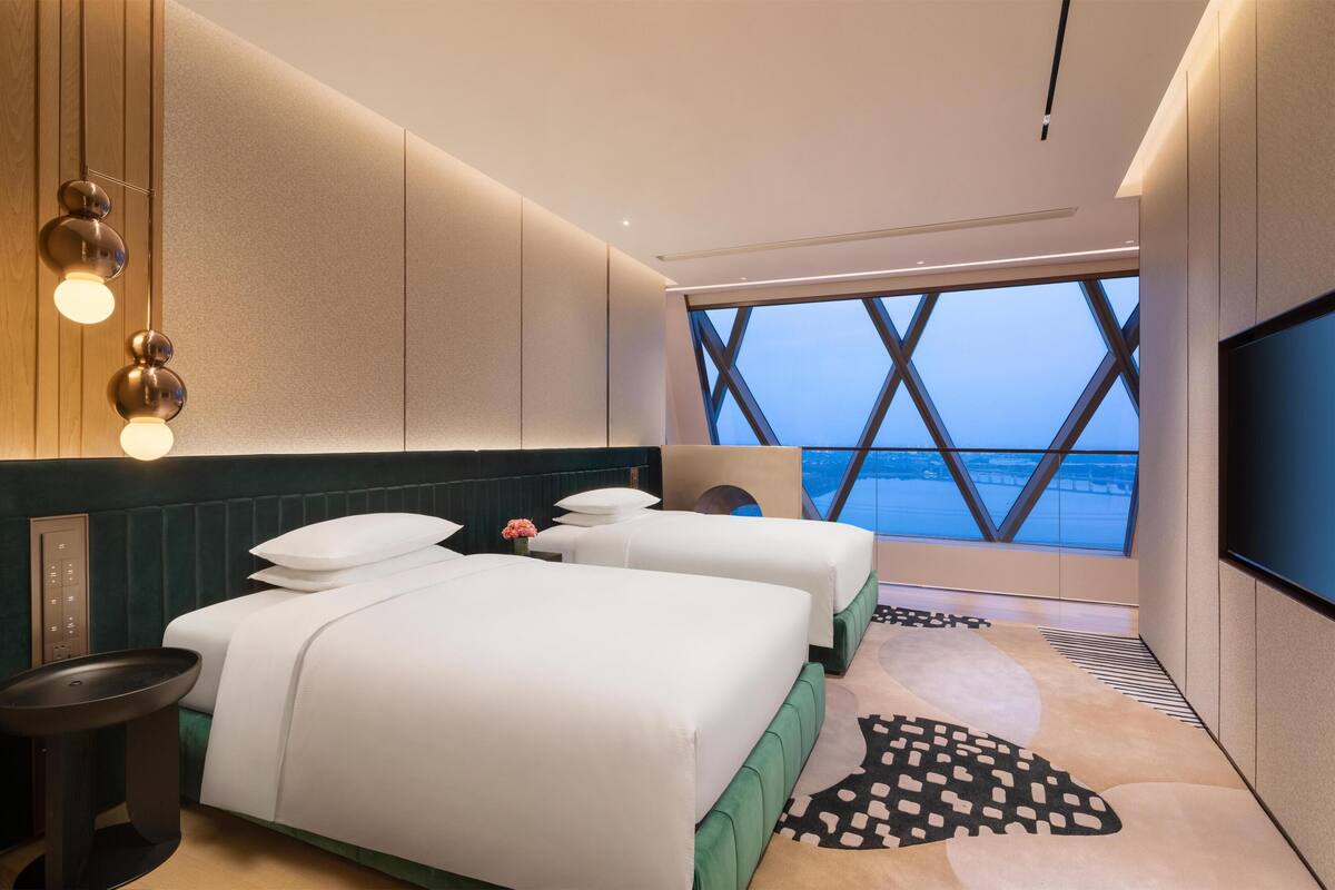 Ein modernes Hotelzimmer mit zwei Betten, einem Fernseher, einem großen Fenster mit Ausblick und einem einzigartigen geometrischen Deckenmuster.