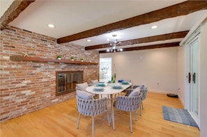 A dining area with a brick wall, wooden beams, and a round table with chairs. - Cape Home w/Oceanview, Roof Deck Pool & Hot Tub (Wellfleet)