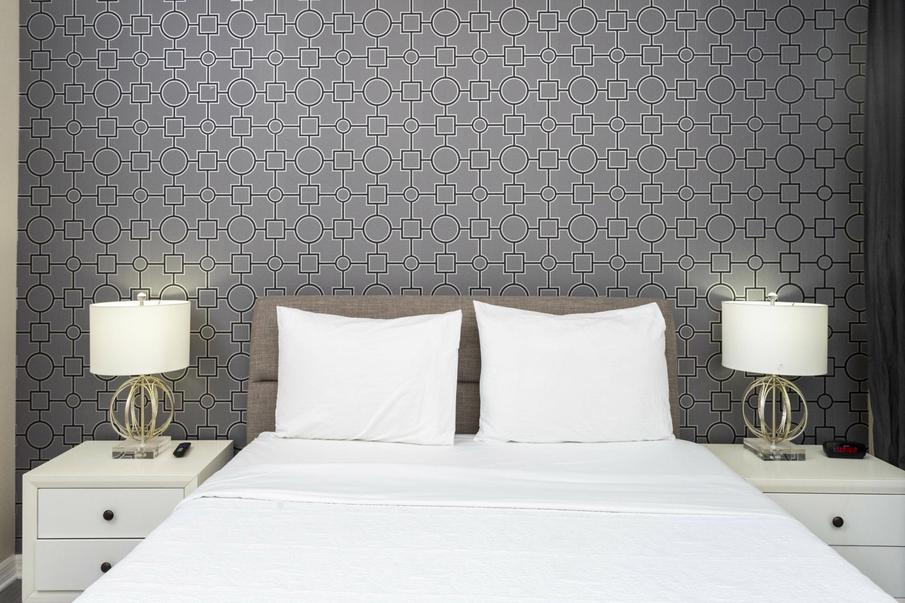 Ein Hotelzimmer mit strukturiertem Wandpapier, einem Bett mit weißer Bettwäsche, zwei Nachttischen mit Lampen und einem Beistelltisch mit einer Uhr.