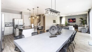 A modern kitchen with white cabinets, stainless steel appliances, and a central island with seating. There are pendant lights above the island and a large flat-screen TV mounted on the wall. - Amazing Dream Family Sleep21 Privatepool Veranda (Kissimmee)