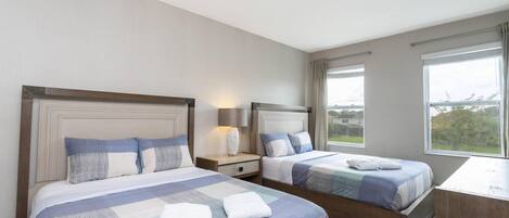 Habitación de hotel con dos camas, una mesita de noche, una lámpara y ventanas amplias con vistas.
