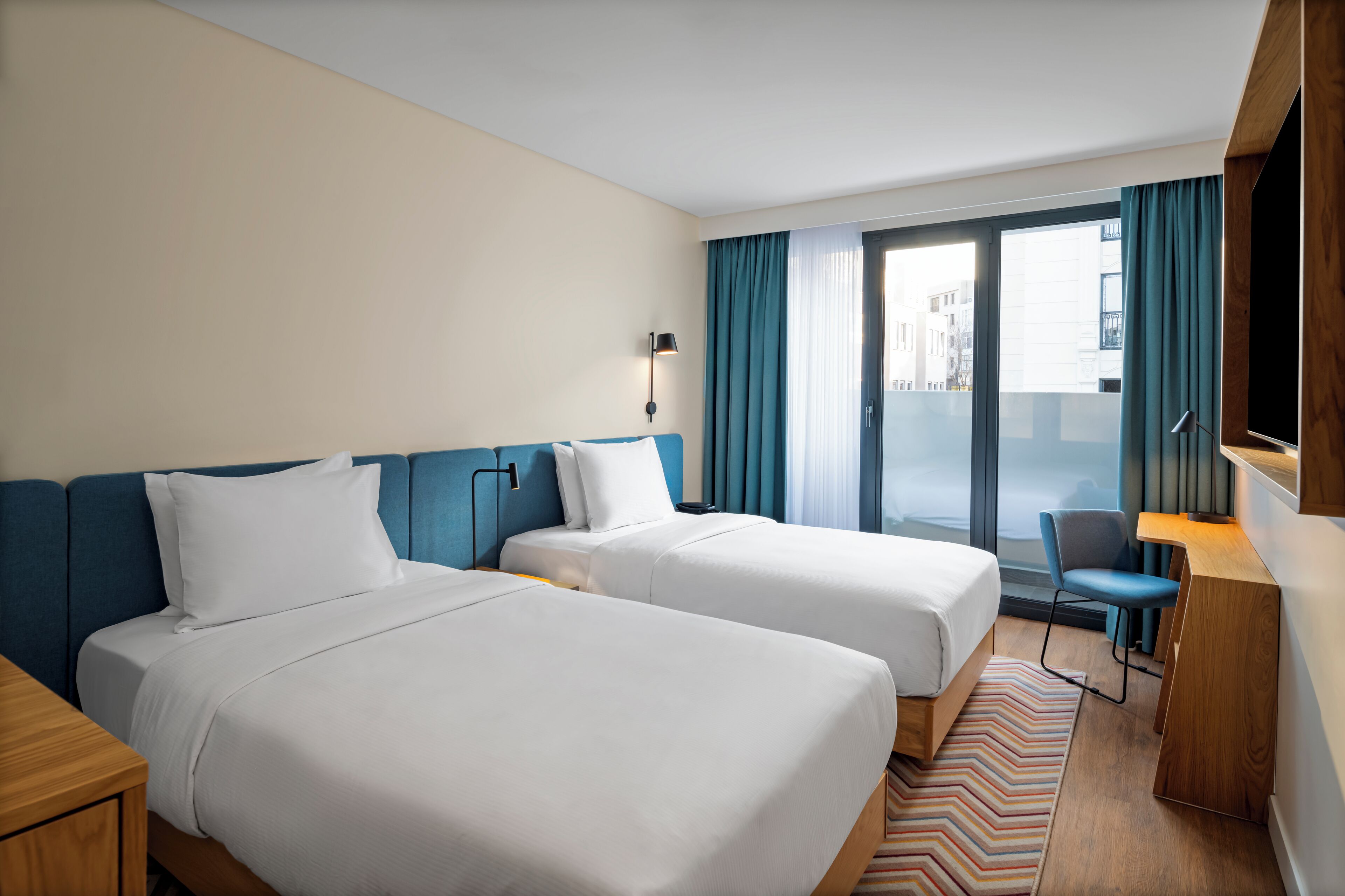 Une chambre d’hôtel avec deux lits, un bureau, une chaise et une grande fenêtre avec des rideaux.