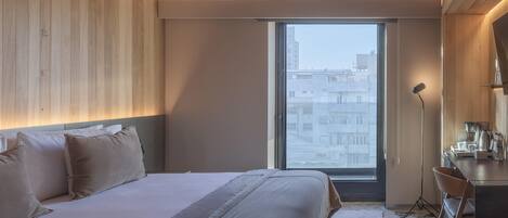 A modern bedroom with a large bed, a desk, and a view of the city through a large window.