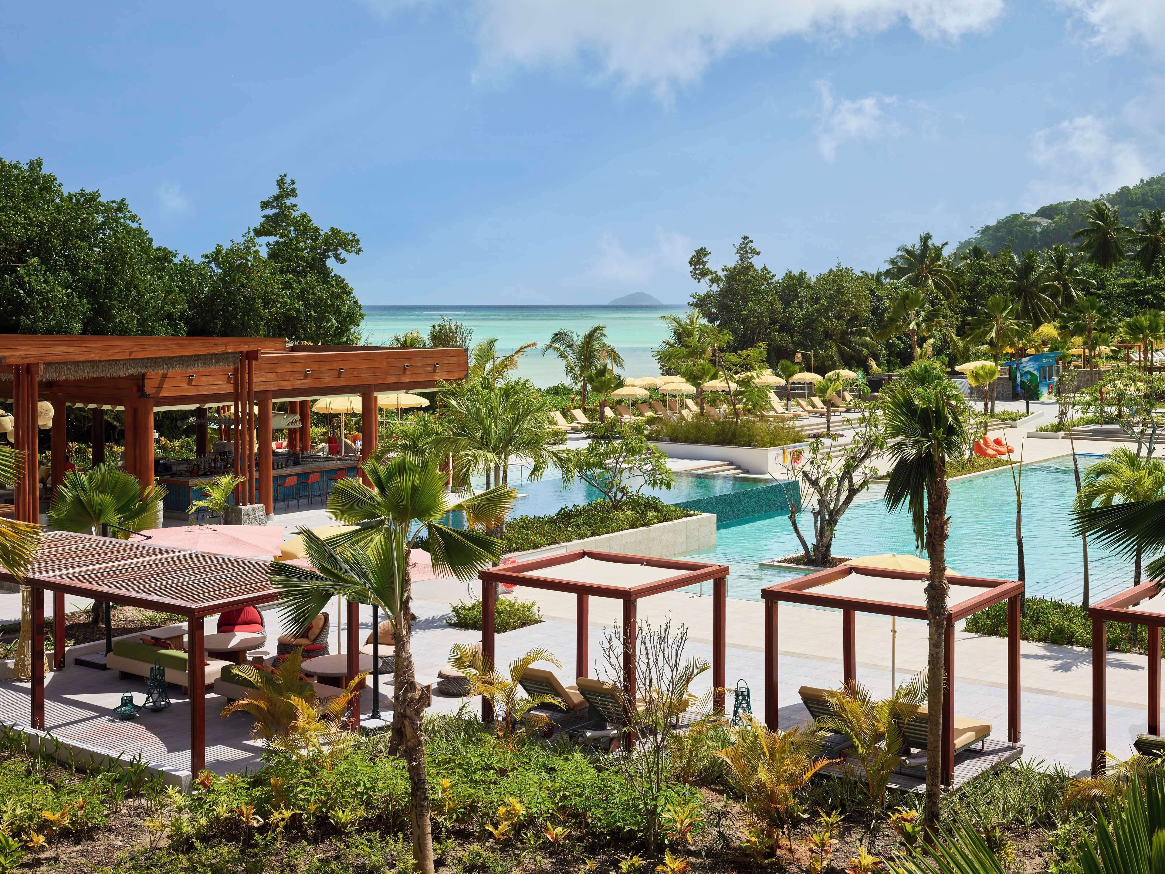 A resort pool area with lounge chairs and a bar, surrounded by tropical plants and overlooking a clear blue sea.