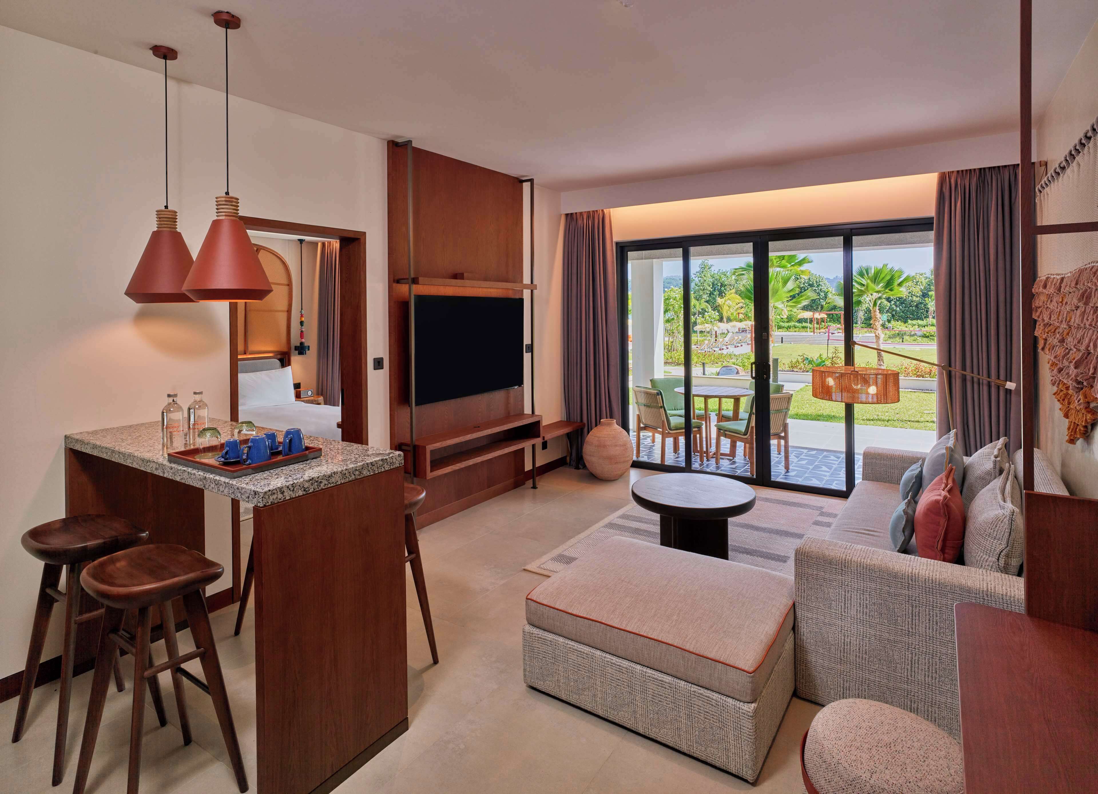 Una habitación de hotel moderna con cocina, zona de estar con sofá y reposapiés, televisor montado en un panel de madera y un amplio ventanal corredero que da a un patio exterior.