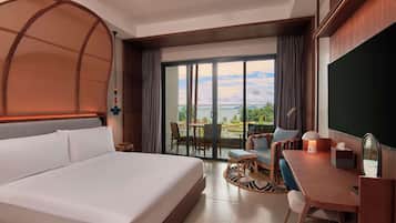 Une chambre d’hôtel avec un grand lit, un bureau avec une chaise, un balcon avec une table et des chaises, et une vue sur la mer.