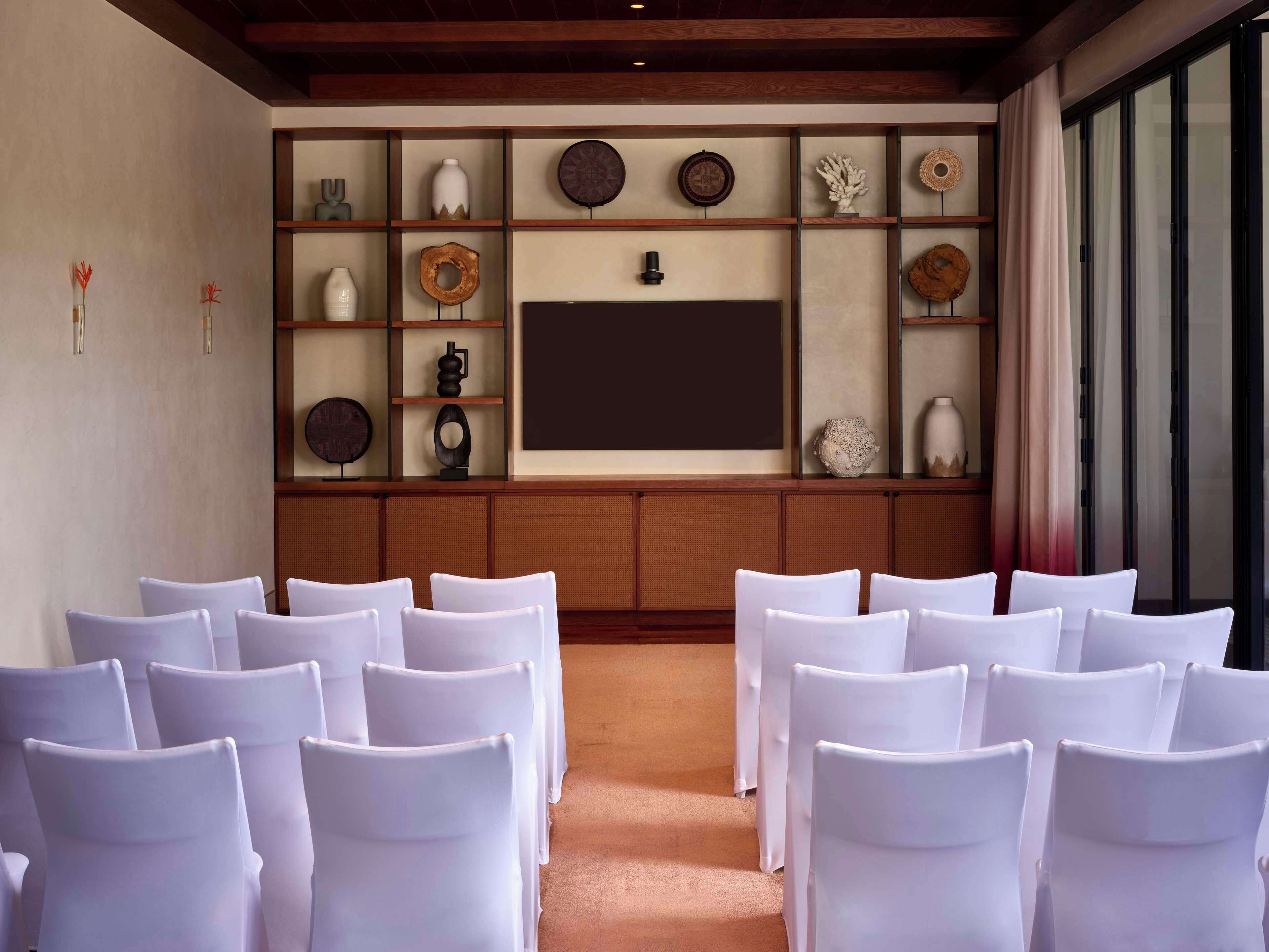 A conference room with white chairs arranged in rows, a large flat-screen TV on the wall, and a wooden shelf with decorative items.