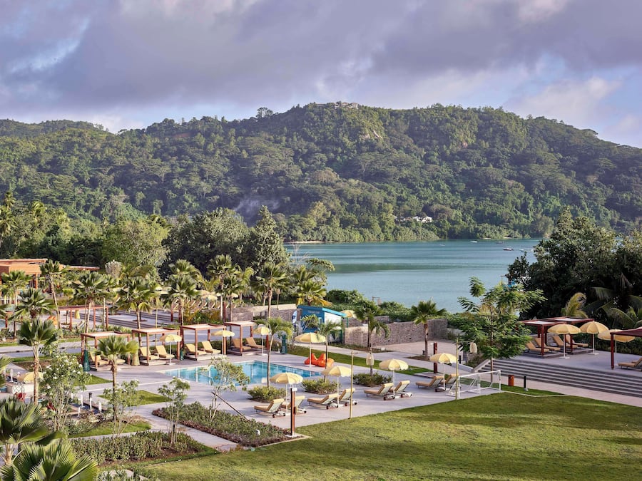 Un resort con piscina, sillas de descanso, sombrillas y vistas a un lago y colinas boscosas.