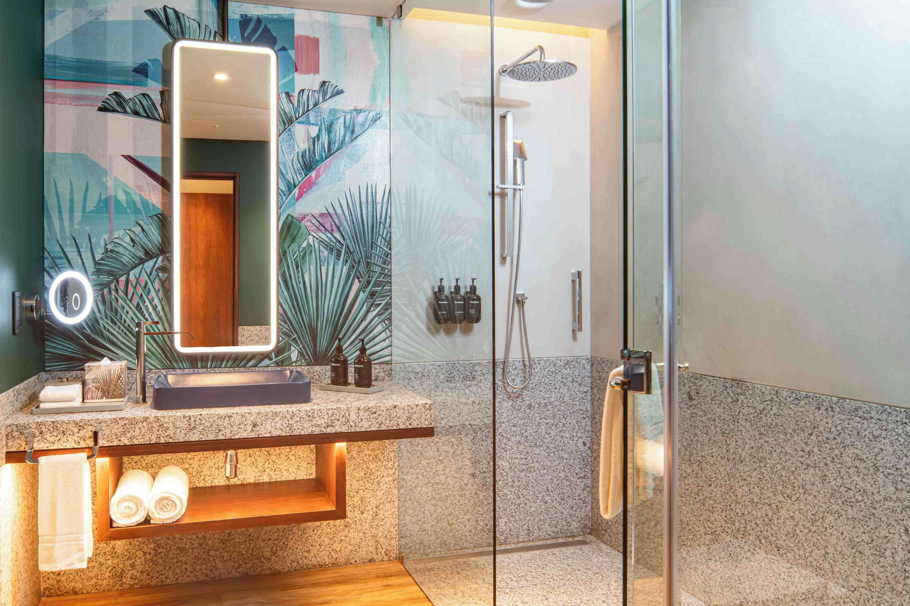 A modern bathroom with a granite countertop, a large mirror with lights, a glass-enclosed shower, and a decorative wall mural.