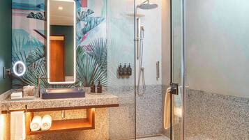 A modern bathroom with a granite countertop, a large mirror with lights, a glass-enclosed shower, and a decorative wall mural.
