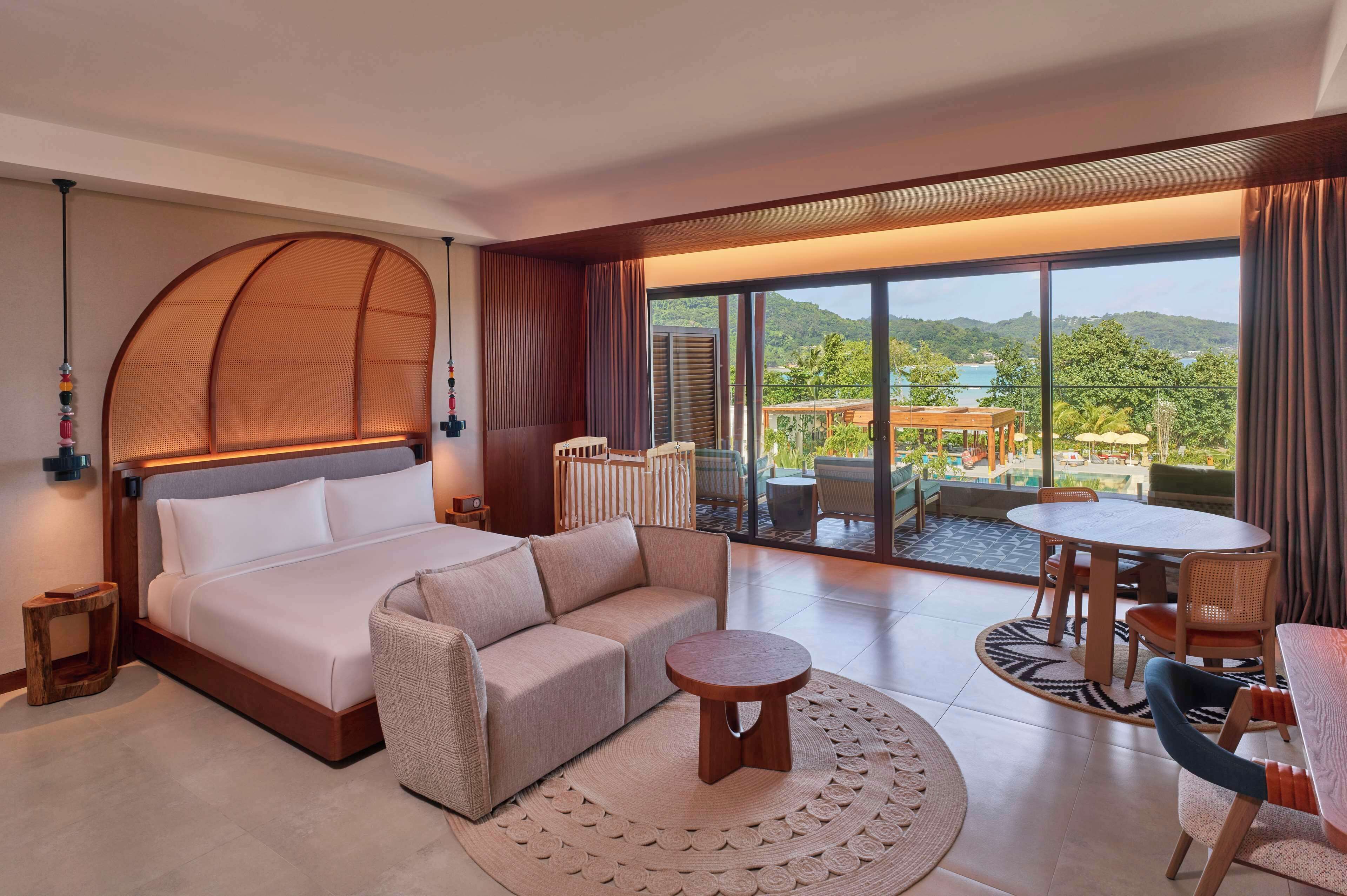 Habitación de hotel con cama, sofá, mesa redonda y balcón con vistas a las montañas y al agua.