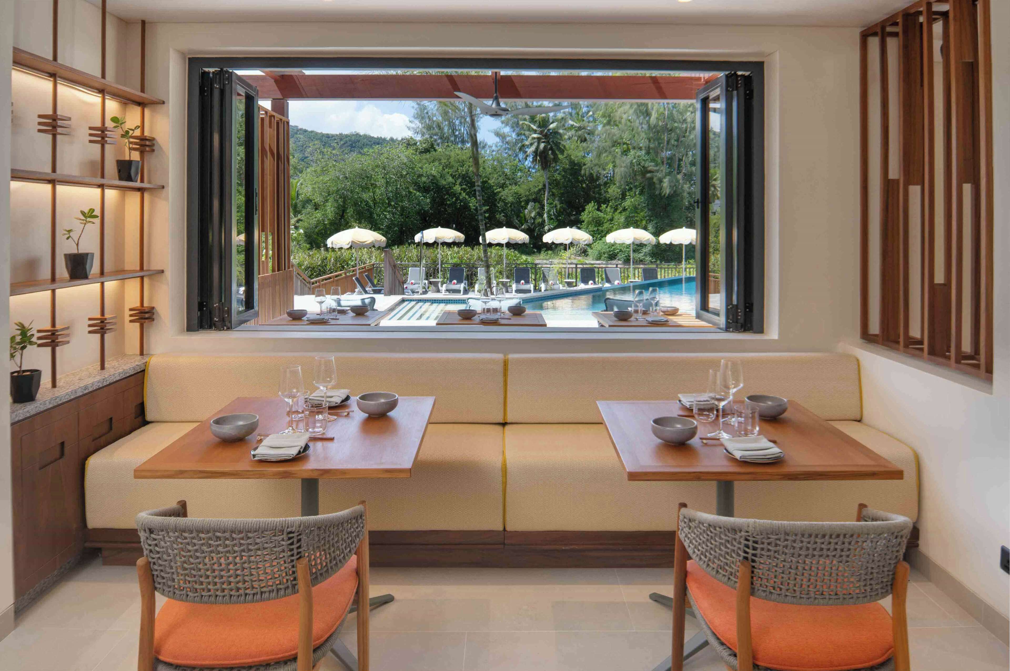 Un espace repas offrant une vue sur une piscine et un jardin luxuriant, comprenant une table en bois, deux bancs rembourrés, des chaises en osier et une étagère avec des plantes en pot.