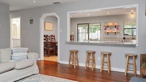 Una sala moderna con un sofá, taburetes de madera y una zona de cocina con ventana y taburetes de bar.