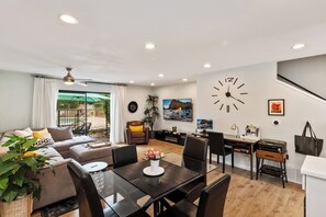 A modern living room with a sofa, dining table, and chairs. There is a large clock on the wall and a staircase leading to another level. - 2BR Poolside - 5 minutes to El Paseo - King Bed - Washer/Dryer (Palm Desert)