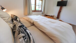 Una camera da letto con un letto, cuscini, un comodino, una lampada, una finestra con tende e una TV a parete.