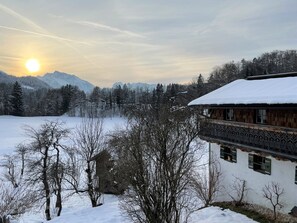 Schneebedeckte Landschaft mit einem Gebäude im traditionellen Stil, umgeben von Bäumen und Bergen im Hintergrund.