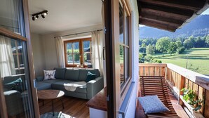 A cozy living room with a sofa and a coffee table, a balcony with wooden railings, and a scenic view of a green landscape.