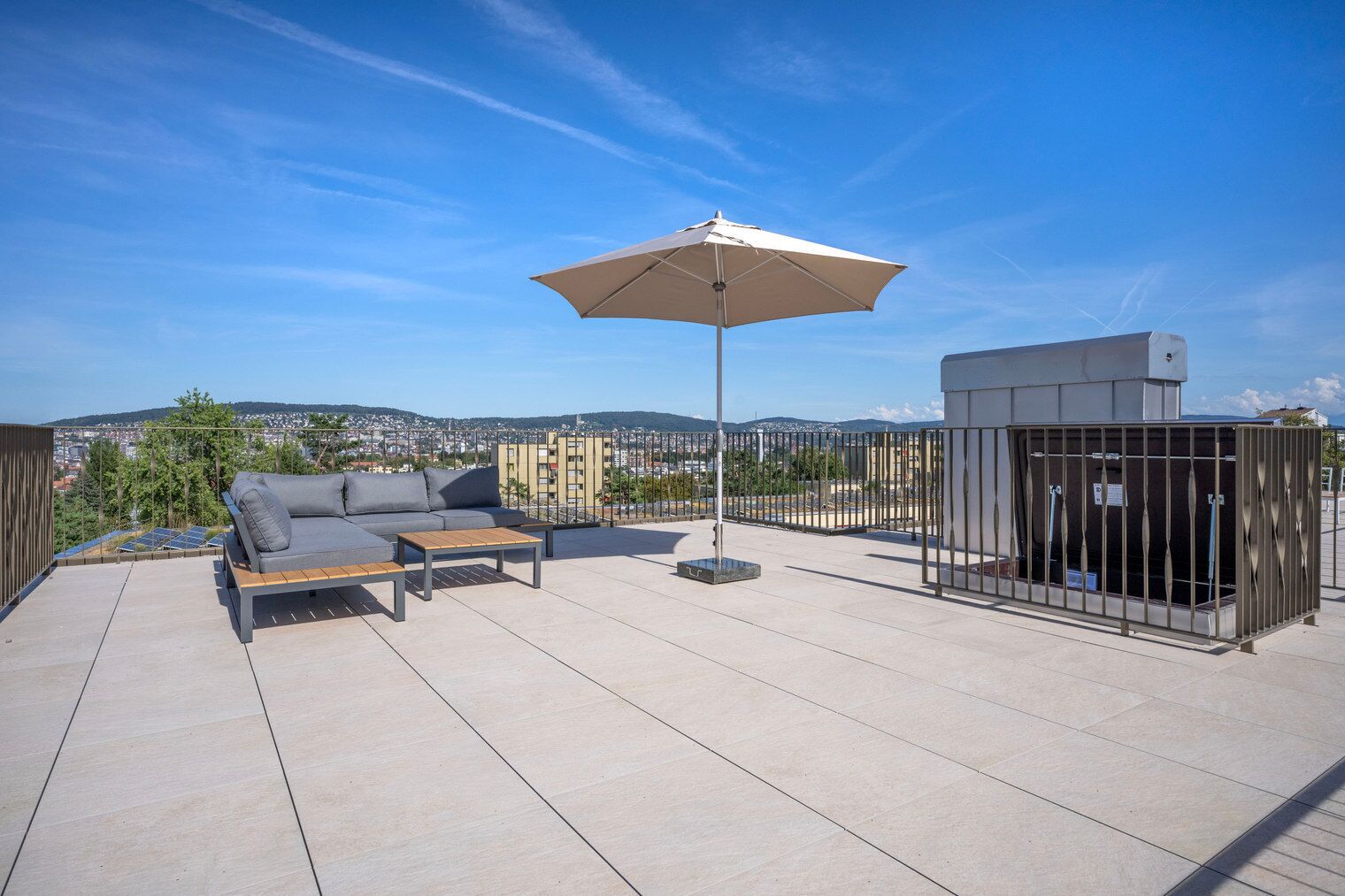 A rooftop terrace with a sofa, a table, and an umbrella. There is a fenced area with a utility box.