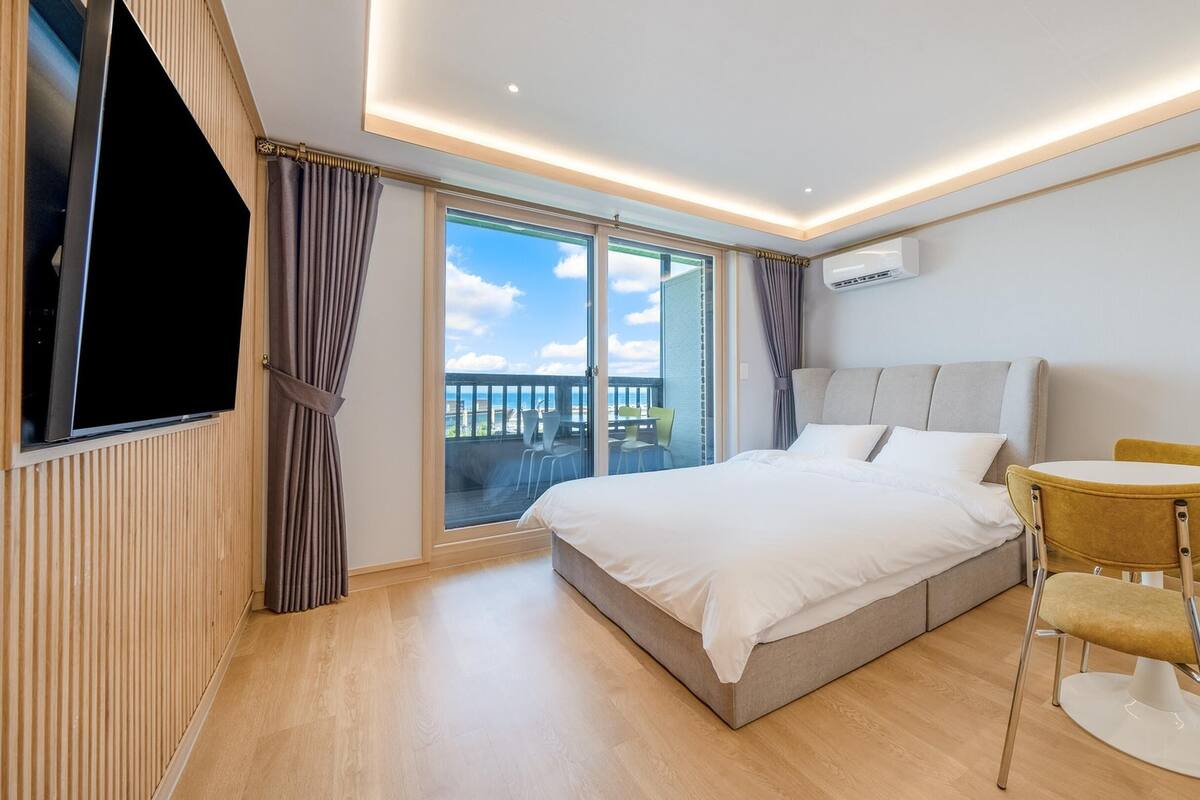 A modern hotel room with a large bed, a TV, a balcony with a view, and a small table with chairs.