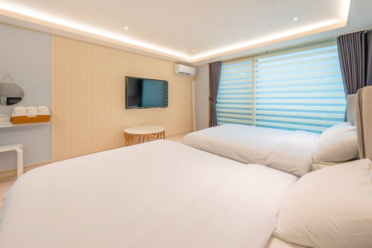A modern bedroom with a large bed, a small round table, a mounted TV, and a window with blinds.