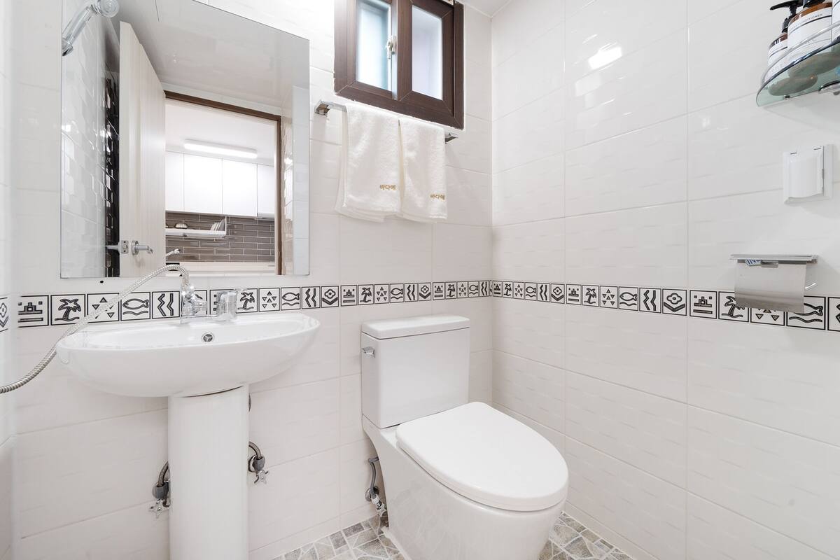 A bathroom with a white sink, toilet, and shower. There is a window above the toilet and a towel rack with towels. The walls are tiled with a border of black and white icons.