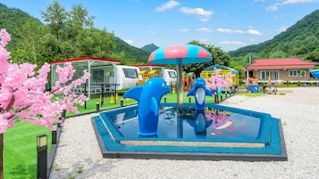 Una piscina con esculturas de delfines azules, rodeada por un área cercada con juegos infantiles y un edificio de techo rojo al fondo.