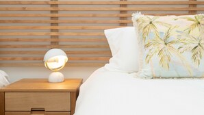 A neatly made bed with a white pillow and a decorative pillow with palm tree patterns. A bedside lamp with a white shade is on a wooden nightstand with a drawer. - Garden View Cottage(2 Bedroom,2 Bathroom) (Kingston)
