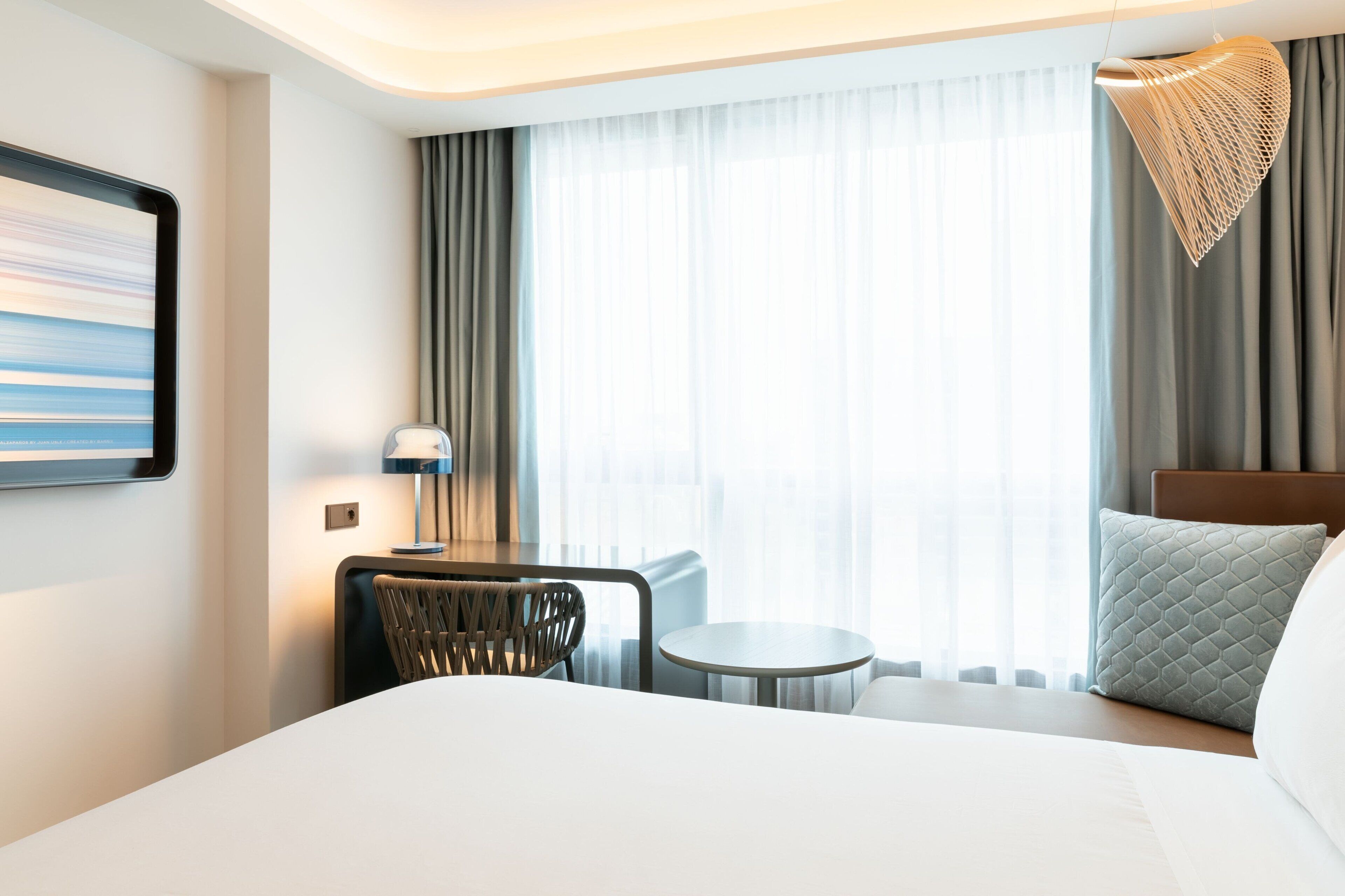 Een hotelkamer met een groot bed, een klein rond tafeltje, een stoel, een lamp en uitzicht op het landschap door dunne gordijnen.