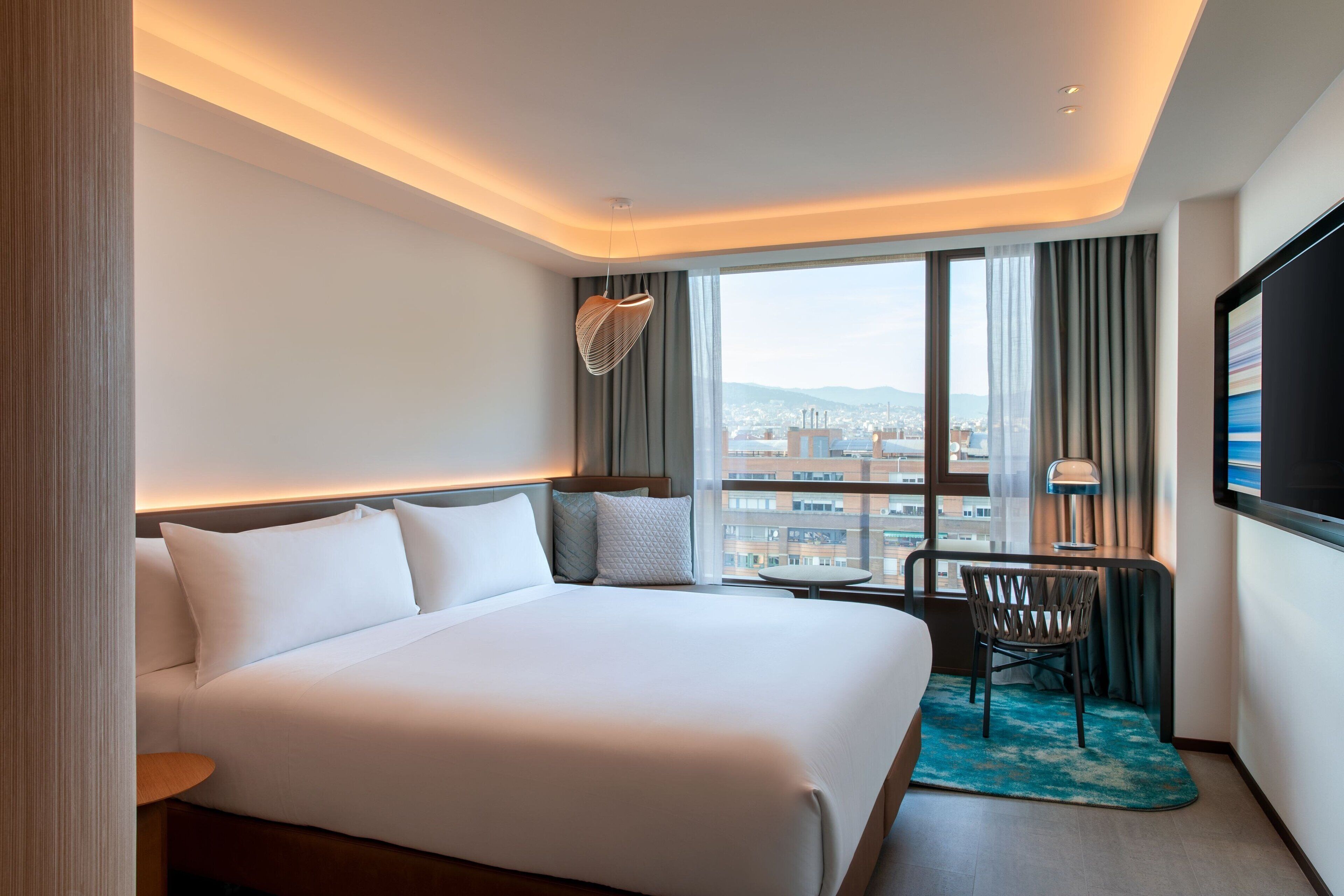 A hotel room with a large bed, a desk with a chair, a TV, and a view of the city.