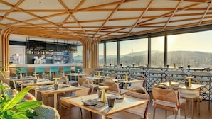 Un restaurant avec un plafond haut, des poutres en bois et une vue sur la ville. Les tables sont dressées avec des assiettes, des couverts et des verres.