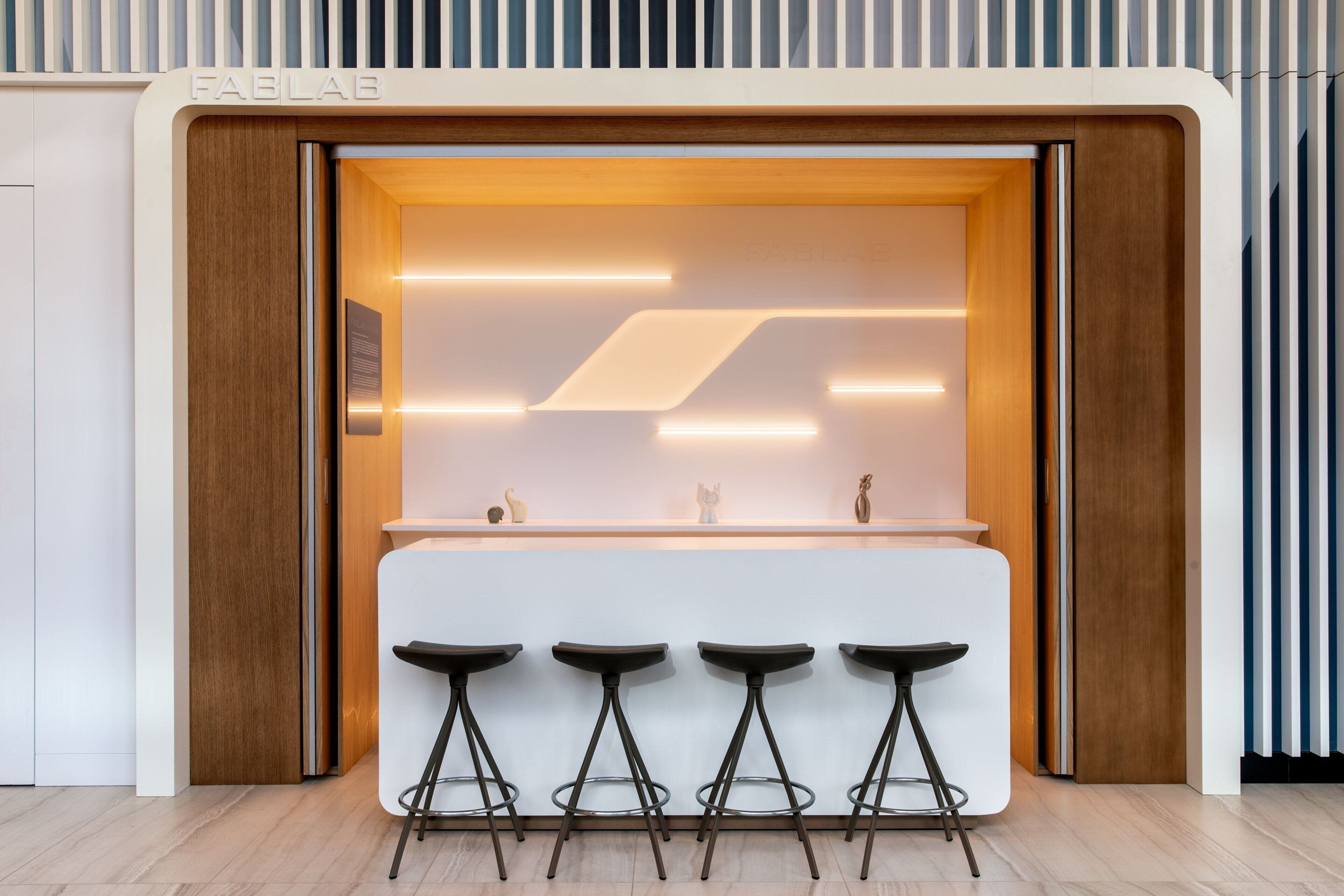 Un área de bar moderna con un revestimiento de pared de madera, una encimera blanca y tres taburetes negros.