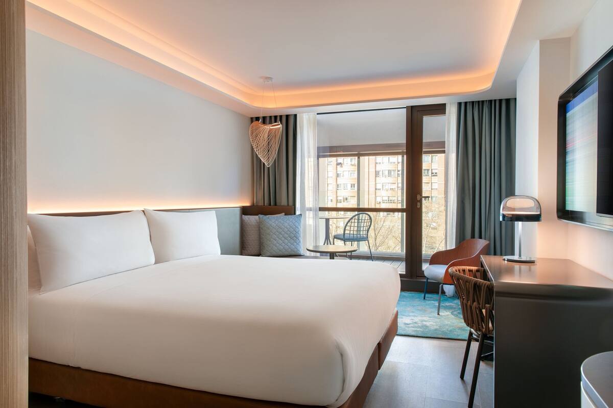 Ein modernes Hotelzimmer mit einem großen Bett, einem Schreibtisch, einem Sessel und Blick auf die Stadt durch das Fenster.