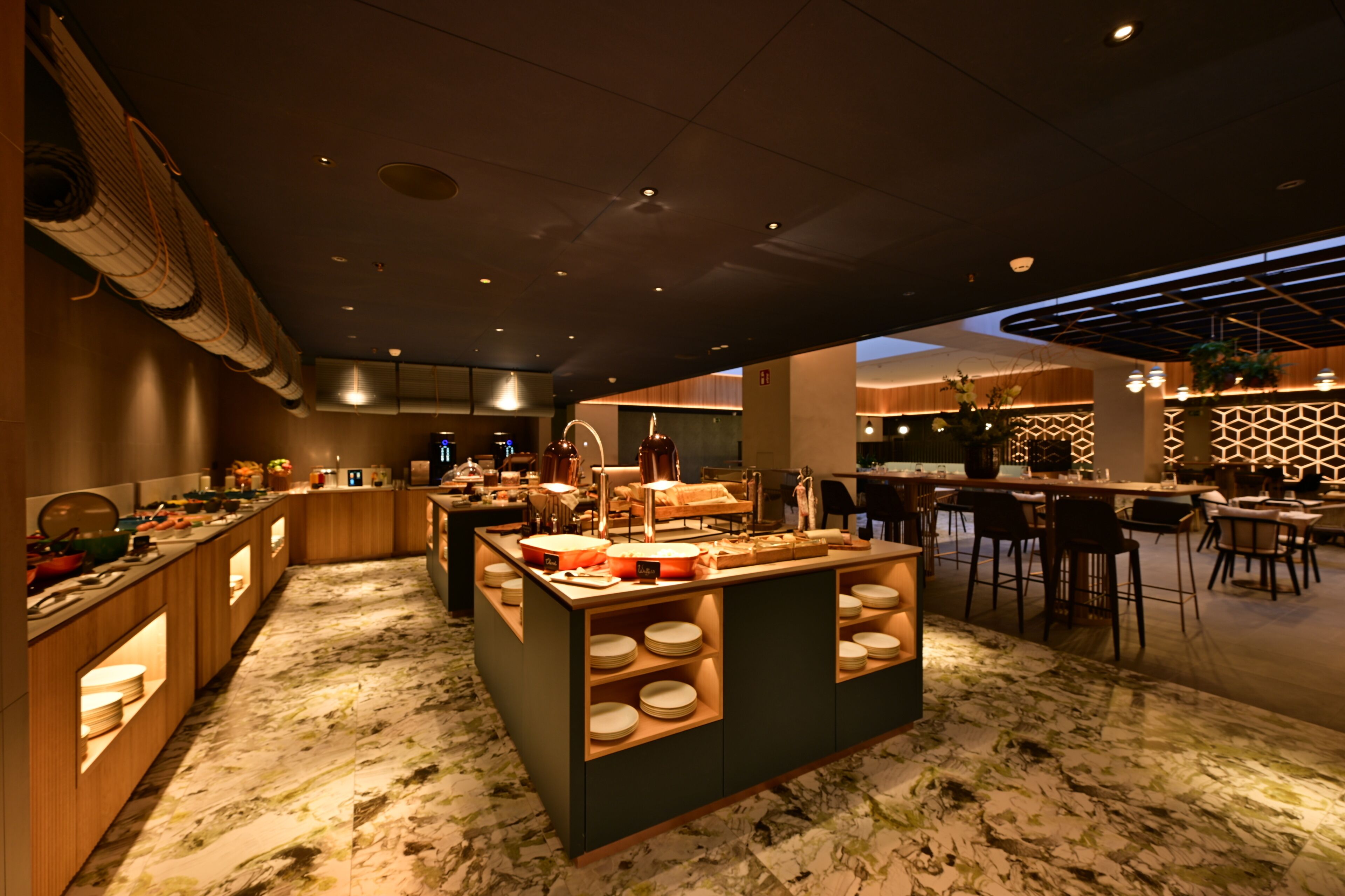 A modern hotel lobby with a central island featuring a buffet, seating areas, and a patterned floor.