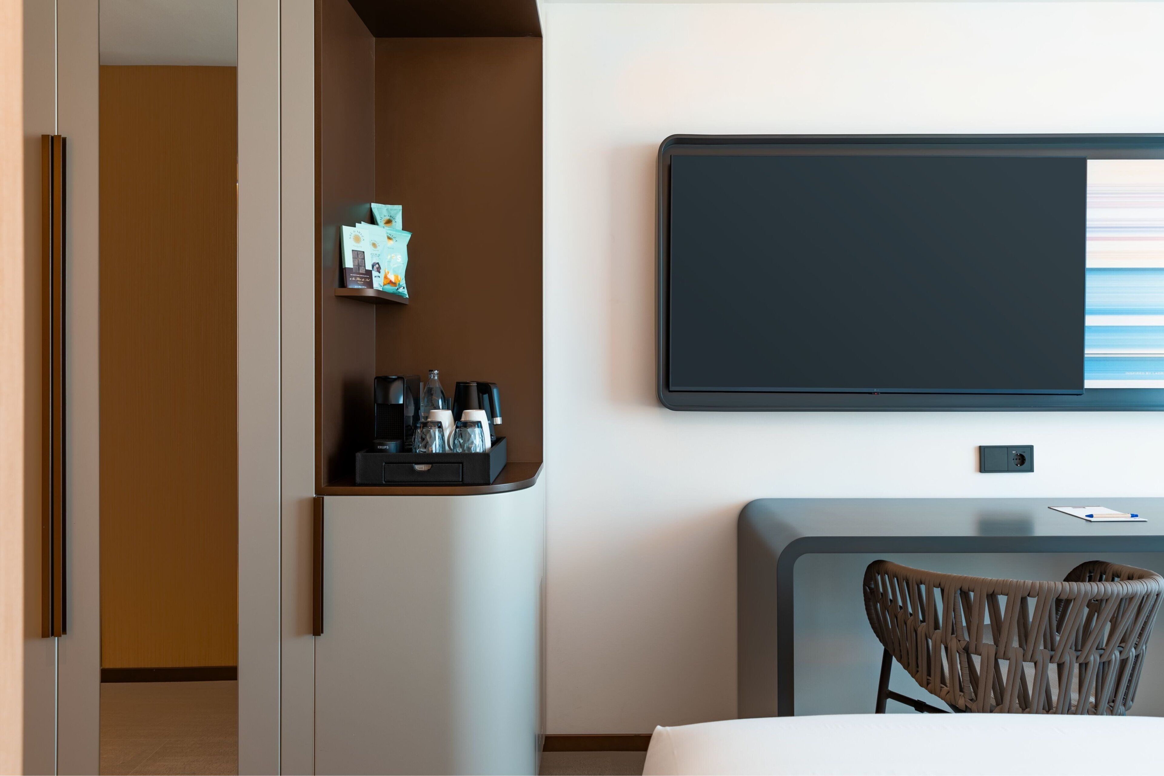 A hotel room with a bed, a desk with a chair, a flat-screen TV mounted on the wall, and a small shelf with a coffee maker and other items.